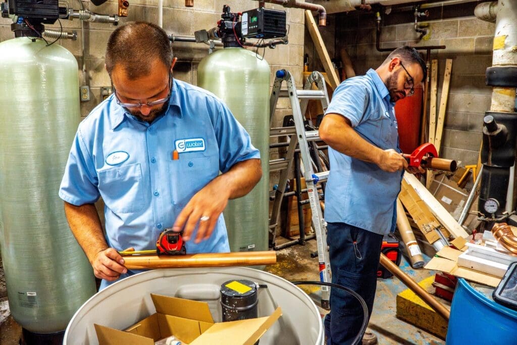 Two water treatment technicians measuring and cutting copper pipe while installing a residential water filtration system